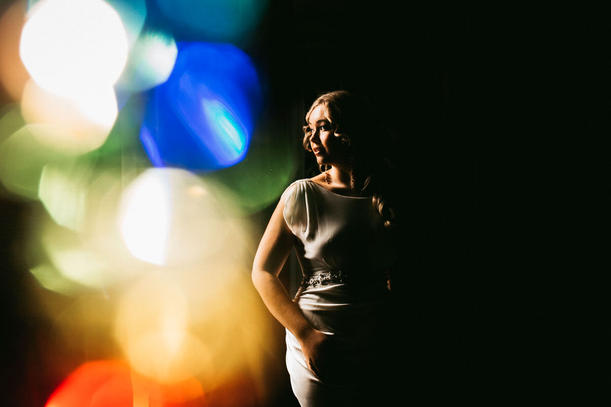 bride looks out of window in a dark room with cool colourful bokeh lighting up 