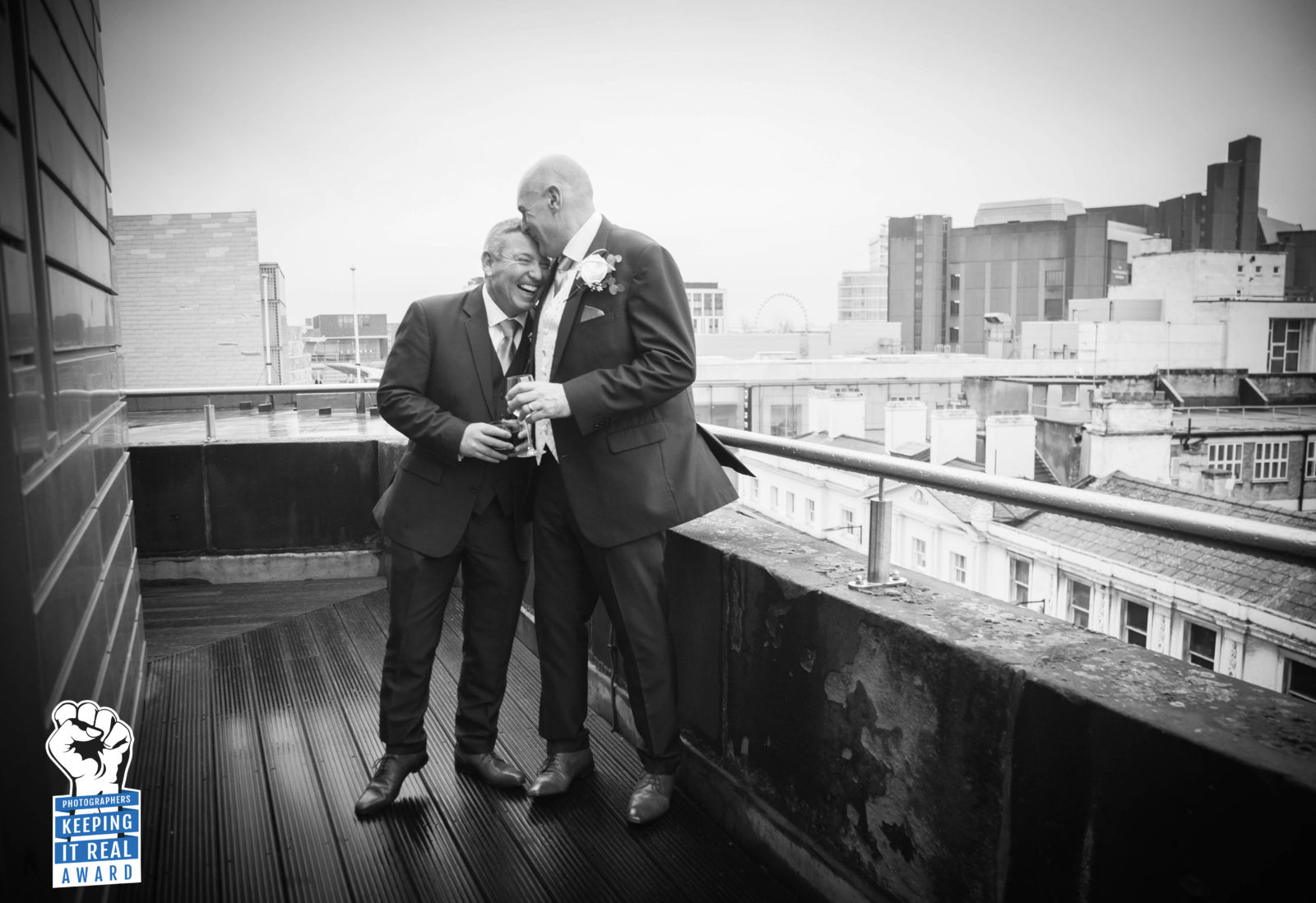 groom embraces his best man as they laugh together about something on the balcony of the john lennon suite looking over liverpool at the hard days night hotel