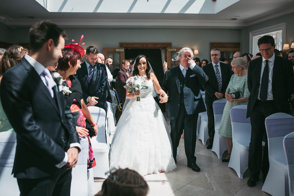 Bride walking down the aisle at willington hall in cheshire looking really happy while her dad wipes a tear from his eye
