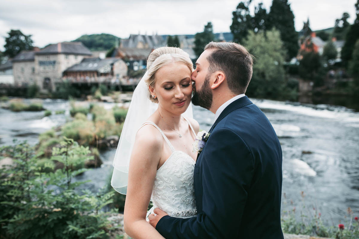 groom kisses his bride in front of the river dee at llangollen railway station